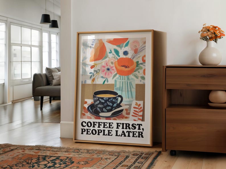 May include: Framed art print featuring a coffee cup with a floral design, a vase of flowers, and the text "COFFEE FIRST, PEOPLE LATER." The artwork has a warm color palette with orange, blue, and brown tones. The print is displayed in a room with a rug and wooden furniture.