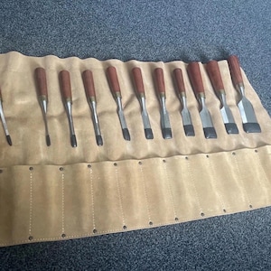 Może przedstawiać: Zestaw dłut do drewna z brązowymi drewnianymi uchwytami i metalowymi ostrzami, ułożonych w beżowym skórzanym rulonie na narzędzia. Dłuta różnią się rozmiarem i kształtem ostrza, do obróbki drewna.