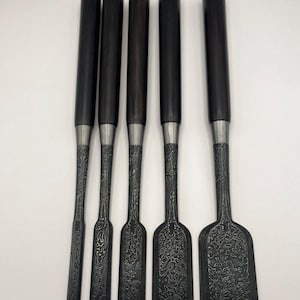 Może przedstawiać: Zestaw pięciu dłut do obróbki drewna. Każde dłuto ma ciemny drewniany uchwyt, srebrną tuleję i wzorzyste, ciemne metalowe ostrze. Ostrza różnią się szerokością, z największym dłutem po prawej stronie. Dłuta są ułożone pionowo na białym tle.