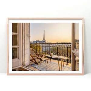 Könnte beinhalten: Ein Balkon mit Blick auf den Eiffelturm in Paris, Frankreich. Der Balkon hat einen Holzstuhl, einen kleinen Tisch mit einem Buch darauf und einen blau-weißen Teppich. Der Balkon ist von einem schmiedeeisernen Geländer umgeben.