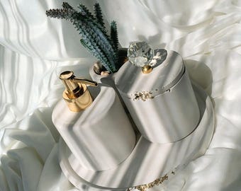 Op de afbeelding: Witte keramische badkamerset met een gouden pompflacon, een pot met deksel met een kristallen knop en een klein vetplantje. De set staat op een witte dienblad met gouden accenten.