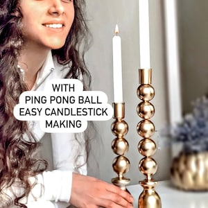 May include: Two gold-colored candlesticks with white candles. The candlesticks have a unique design, featuring stacked spheres. The image includes the text "WITH PING PONG BALL EASY CANDLESTICK MAKING".