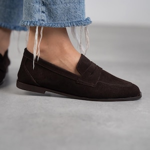 May include: Brown suede leather loafers with a penny strap detail. The loafers have a rounded toe and a low heel.