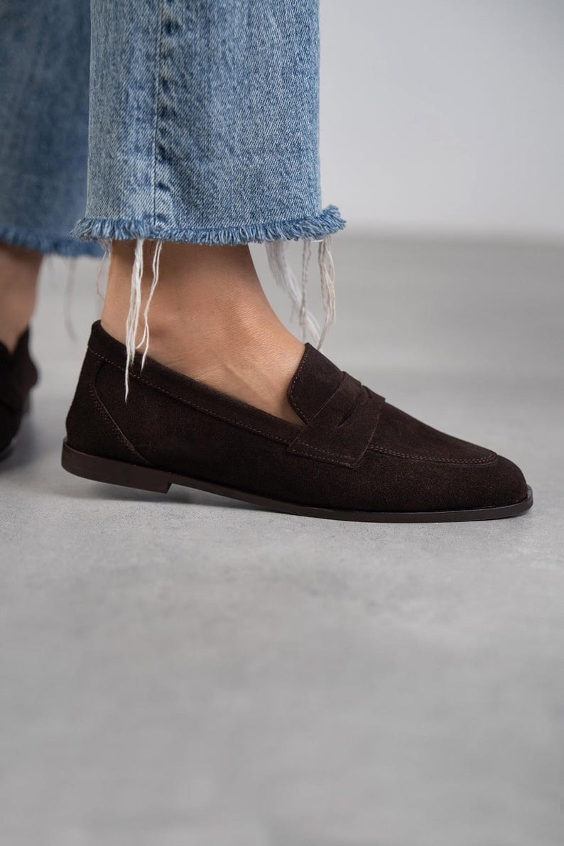 May include: Brown suede leather loafers with a penny strap detail. The loafers have a rounded toe and a low heel.