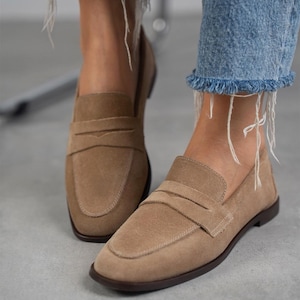 May include: A pair of light brown suede loafers with a buckle detail. The loafers have a rounded toe and a low heel.
