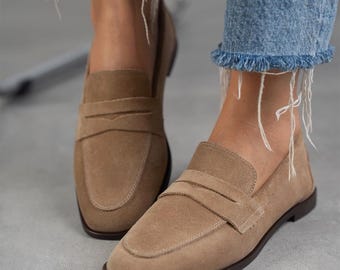 May include: A pair of light brown suede loafers with a buckle detail. The loafers have a rounded toe and a low heel.