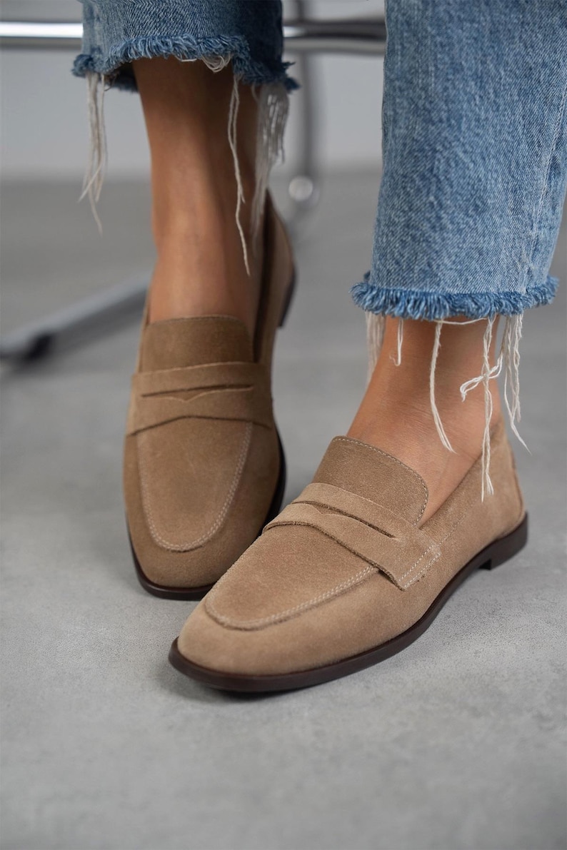 May include: A pair of light brown suede loafers with a buckle detail. The loafers have a rounded toe and a low heel.