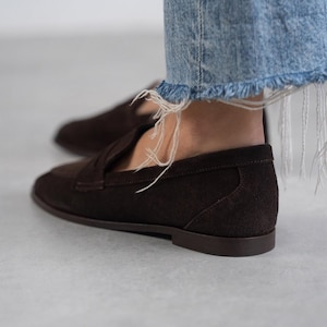 May include: Brown suede loafers with a tassel detail. The loafers have a rounded toe and a low heel.
