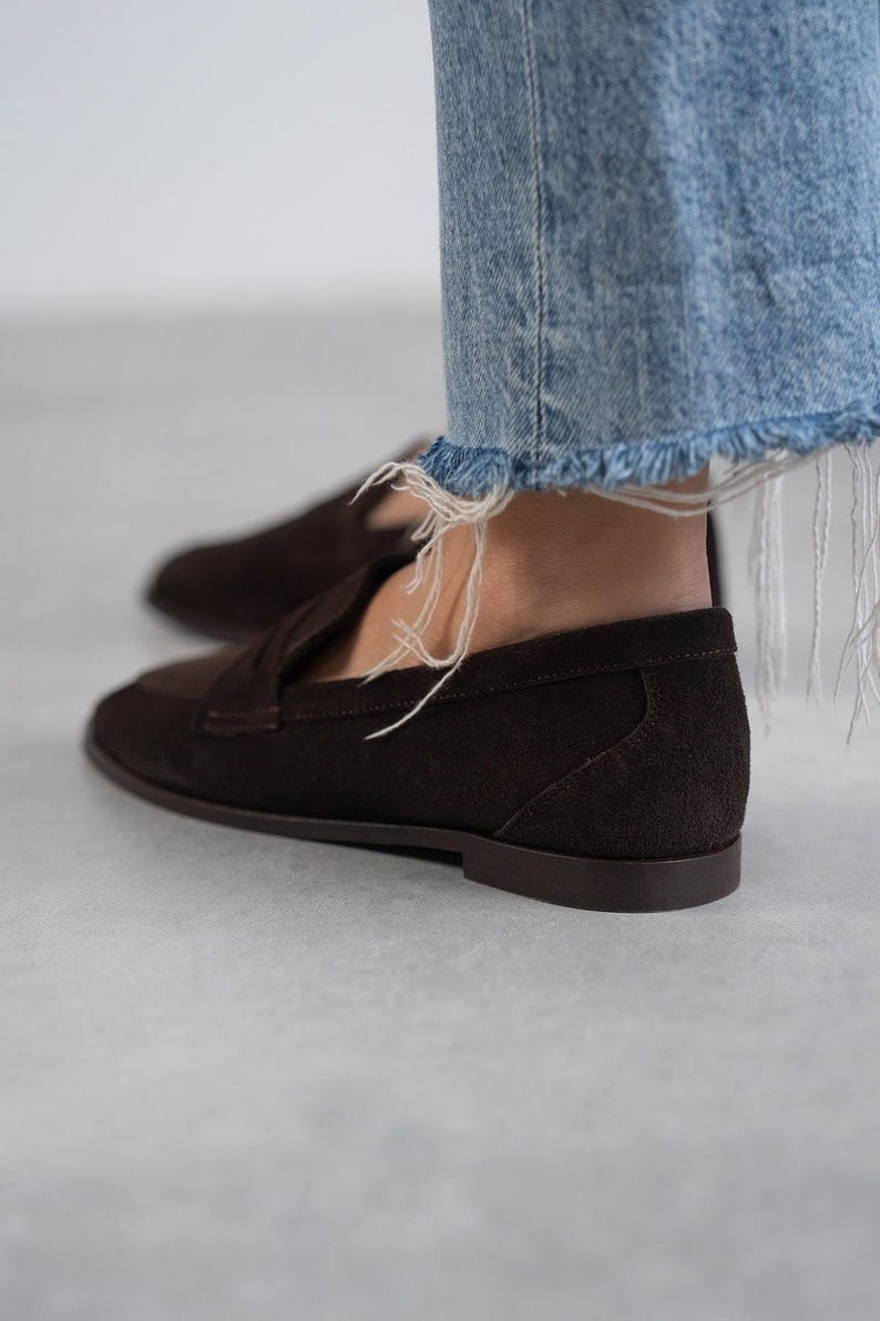 May include: Brown suede loafers with a tassel detail. The loafers have a rounded toe and a low heel.
