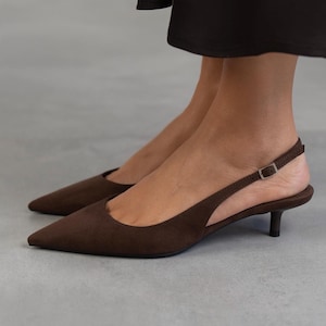 Puede incluir: Zapatos de tacón de gamuza marrón con tira trasera y punta puntiaguda.