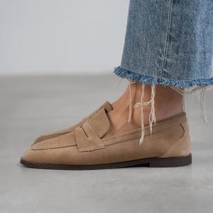 May include: A pair of light brown suede loafers with a simple design. The loafers have a rounded toe and a low heel.