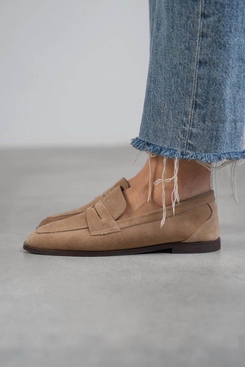May include: A pair of light brown suede loafers with a simple design. The loafers have a rounded toe and a low heel.