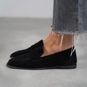 May include: Black suede loafers with a classic penny strap design. The loafers have a rounded toe and a low heel.