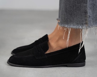 May include: Black suede loafers with a classic penny strap design. The loafers have a rounded toe and a low heel.