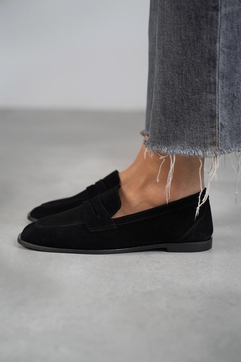 May include: Black suede loafers with a classic penny strap design. The loafers have a rounded toe and a low heel.