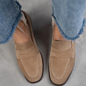 May include: A pair of light brown suede loafers with a simple design. The loafers have a rounded toe and a low heel.
