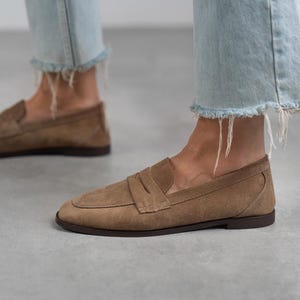 May include: A pair of brown suede loafers with a simple design. The loafers have a rounded toe and a low heel.