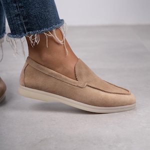 May include: Tan suede slip-on loafers with a light-colored sole. The shoes have a classic design with a rounded toe and a low heel. The person is wearing blue jeans with frayed edges.