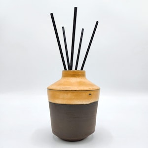 Może przedstawiać: Ceramiczna butelka dyfuzora z brązową i żółtą glazurą. Butelka ma czarne patyczki dyfuzora włożone do górnej części.