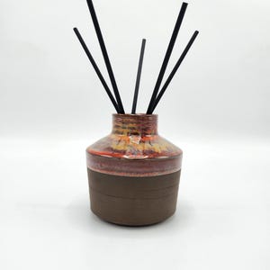 May include: A ceramic essential oil diffuser with a brown base and a reddish-orange top. The diffuser holds several black reeds, creating a decorative and aromatic display. The design is simple and elegant.