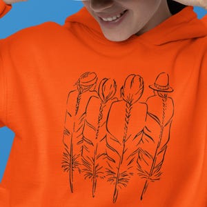 May include: An orange hoodie featuring a black line drawing of four figures with long braids. A white baseball cap is being held up by a person with red nail polish. The background is a solid blue.