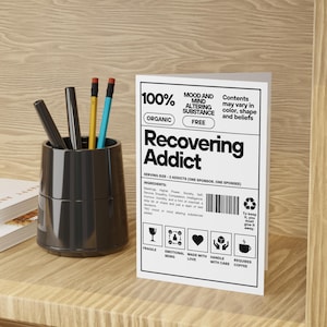 May include: A white greeting card with the text "Recovering Addict" in bold black letters. The card includes a list of ingredients and icons. A black pen holder with pens and pencils sits to the left of the card.