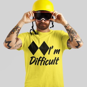 May include: A yellow t-shirt with the words "I'm Difficult" in black script, and two black diamond ski slope symbols. The person is wearing a yellow ski helmet and goggles.