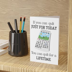 Può includere: Un biglietto di auguri bianco con il testo "If you can quit JUST FOR TODAY You can quit for a LIFETIME." La carta presenta un'illustrazione di calendario con una scena di montagna. Un portapenne nero con penne e matite è a sinistra.