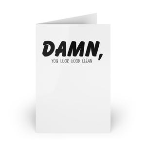 May include: A white greeting card with the text "Damn, You Look Good Clean" in a black, bold, handwritten font. The card is folded and stands upright, featuring a simple, minimalist design.