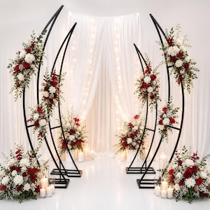 Könnte beinhalten: Zwei schwarze, halbmondförmige Metallbögen, die mit Arrangements aus roten und weißen Blumen sowie grünem Blattwerk geschmückt sind. Die Bögen stehen auf einem weißen Boden, ergänzt durch weiße Stumpenkerzen und Blumensträuße am Fuß.