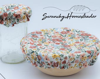May include: Two fabric bowl covers with an elasticized edge, featuring a white background with a floral pattern of red, yellow, and green leaves. One cover is on a glass jar, and the other is on a round bowl. Text reads Scrunchy Homesteader.