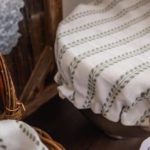 May include: A round fabric bowl cover featuring a white base with vertical stripes of small, sage green leaf patterns. The cover is secured with an elastic edge and a piece of twine tied in a small bow, resting over a bowl with a visible tag.