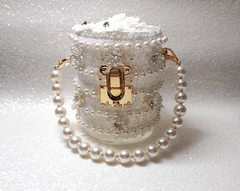 Puede incluir: Un bolso blanco cilíndrico con una correa de perlas y un cierre dorado. El bolso está cubierto de encaje blanco y perlas.