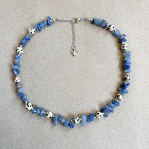 May include: A blue stone necklace with Dalmatian jasper beads. The necklace features small, irregular blue stones interspersed with round beads with black spots. The necklace has a silver-coloured chain and clasp, with a small charm.