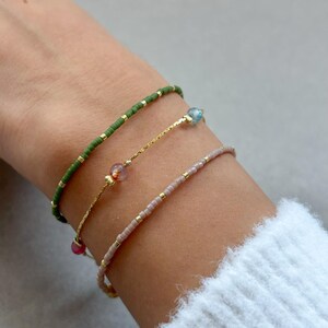 Könnte beinhalten: Drei zarte Armbänder mit kleinen Perlen. Das obere Armband ist grün, das mittlere Armband hat eine goldene Kette mit einem rosa Stein und das untere Armband ist beige.