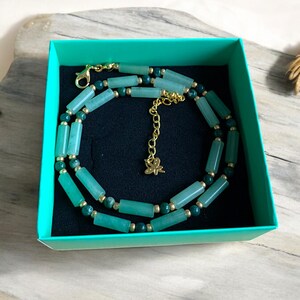 Pode incluir: Um colar de jade verde com detalhes dourados em uma caixa de presente turquesa. O colar tem um pequeno pingente de borboleta dourado.