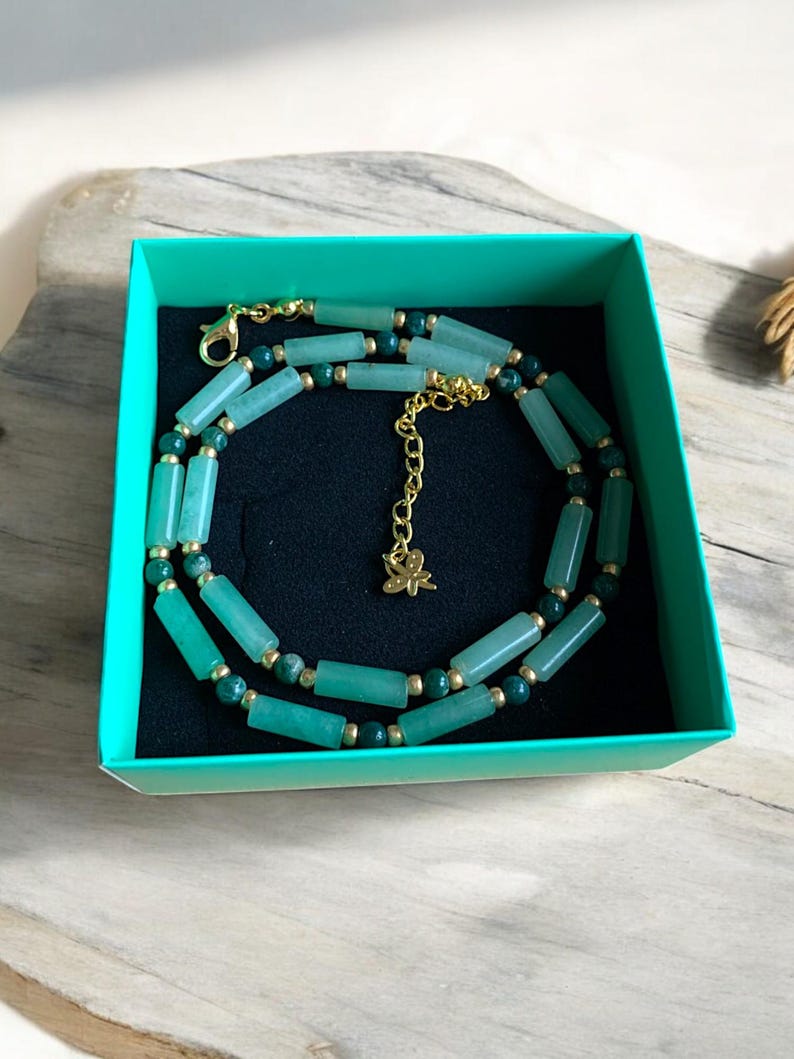 Pode incluir: Um colar de jade verde com detalhes dourados em uma caixa de presente turquesa. O colar tem um pequeno pingente de borboleta dourado.