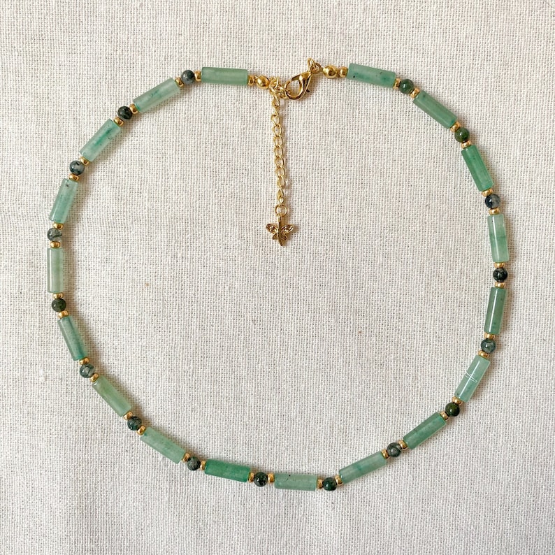 Pode incluir: Um colar de contas verdes e douradas com um pequeno pingente de borboleta. O colar &eacute; feito de contas de pedra verde e espa&ccedil;adores dourados.