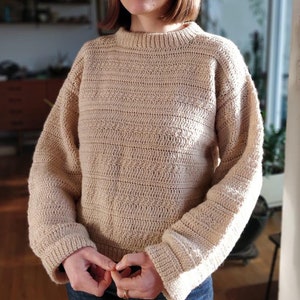 Può includere: Un maglione beige lavorato a uncinetto con scollo rotondo e maniche lunghe. Il maglione ha un motivo a punto testurizzato.