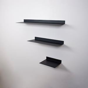 May include: Three black metal floating shelves of varying sizes are mounted on a white wall. The shelves have a simple, modern design with a flat surface and a raised back edge. The shelves are arranged vertically, creating a minimalist display.