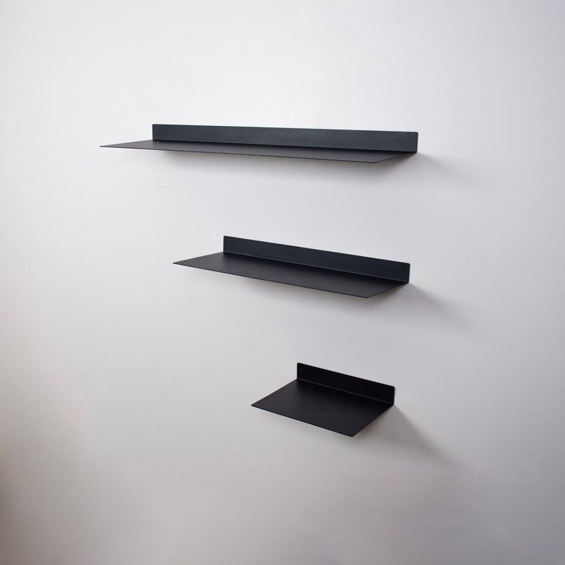 May include: Three black metal floating shelves of varying sizes are mounted on a white wall. The shelves have a simple, modern design with a flat surface and a raised back edge. The shelves are arranged vertically, creating a minimalist display.