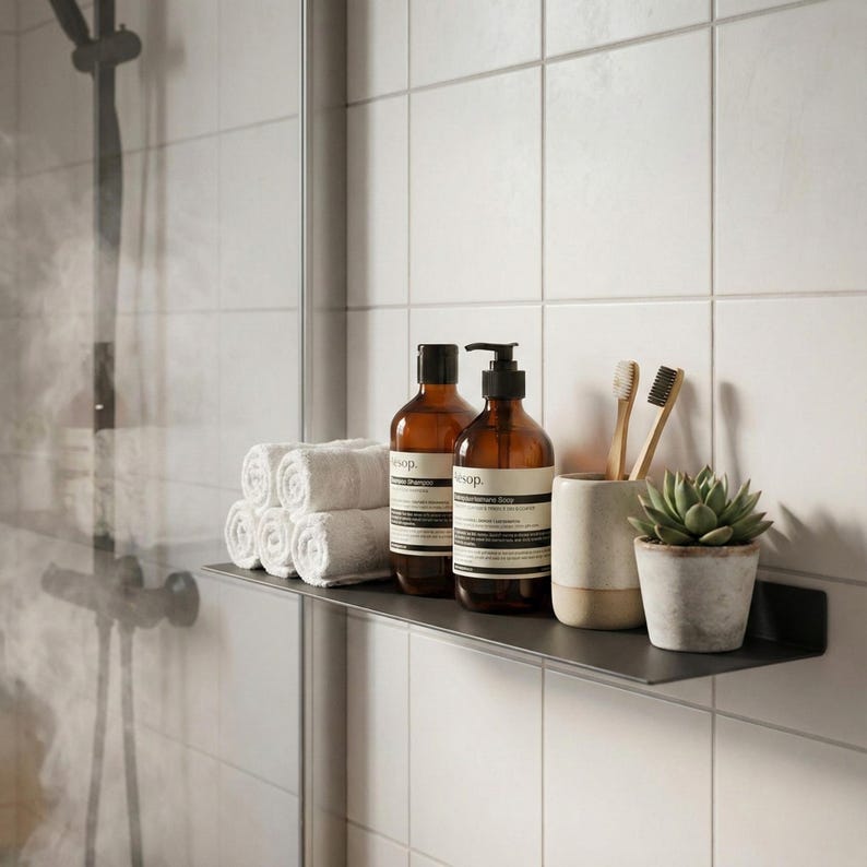 May include: A bathroom shelf displays rolled white towels, brown glass bottles of soap and shampoo, a ceramic cup with toothbrushes, and a small potted succulent. The shelf is mounted on a white tiled wall.