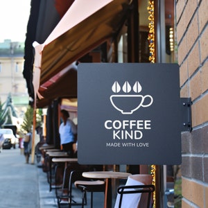 May include: A black sign with white text and a coffee cup logo reads "Coffee Kind, Made with Love". The sign is mounted on a brick wall outside a cafe with tables and chairs.