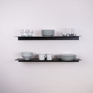May include: Two black floating shelves mounted on a white wall, each holding dishware. The top shelf displays clear drinking glasses, gray plates, and a stack of gray plates. The bottom shelf holds gray bowls, white mugs, and gray plates.