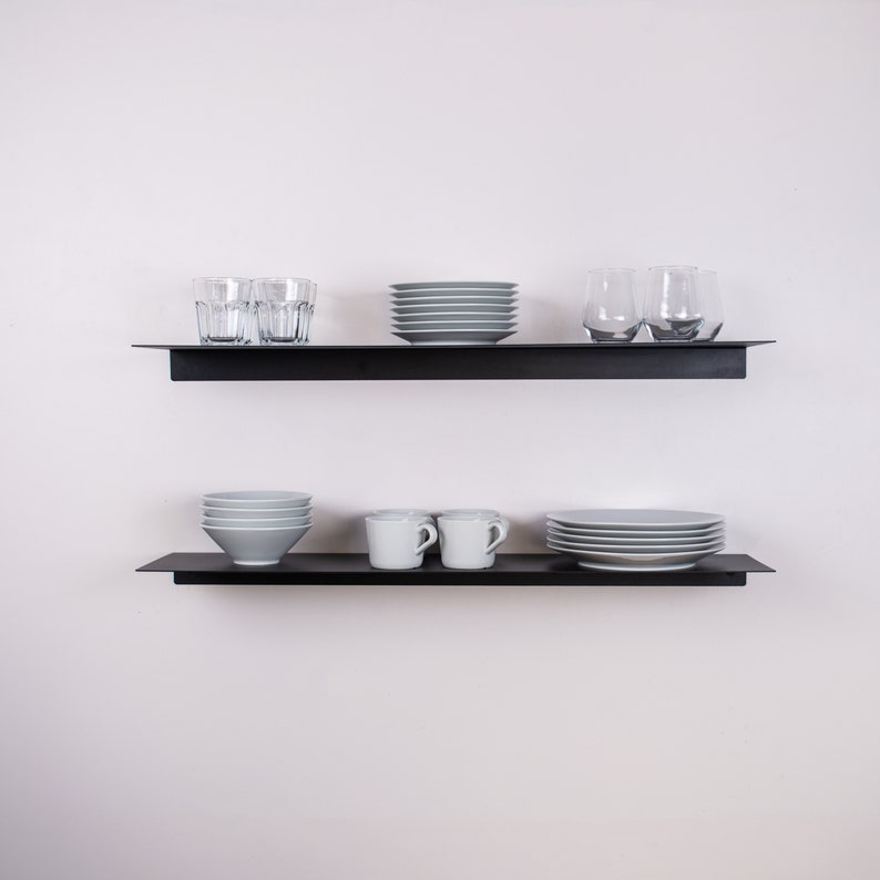May include: Two black floating shelves mounted on a white wall, each holding dishware. The top shelf displays clear drinking glasses, gray plates, and a stack of gray plates. The bottom shelf holds gray bowls, white mugs, and gray plates.