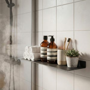 May include: A bathroom shelf displays rolled white towels, brown glass bottles of soap and shampoo, a ceramic cup with toothbrushes, and a small potted succulent. The shelf is mounted on a white tiled wall.