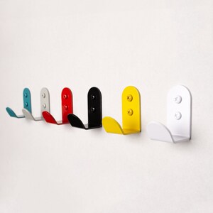Puede incluir: Seis ganchos de metal de diferentes colores, incluyendo azul, gris, rojo, negro, amarillo y blanco, montados en una pared blanca. Los ganchos tienen todos la forma de un gancho simple y redondeado.