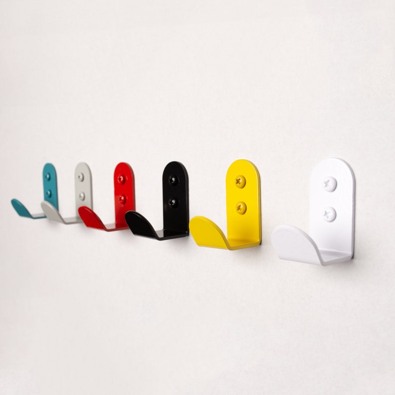 Puede incluir: Seis ganchos de metal de diferentes colores, incluyendo azul, gris, rojo, negro, amarillo y blanco, montados en una pared blanca. Los ganchos tienen todos la forma de un gancho simple y redondeado.