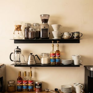 May include: A coffee station with various items on black shelves. Includes a coffee grinder, glass jars with coffee beans, ceramic mugs, a French press, a kettle, Torani vanilla and caramel syrups, and espresso powder.