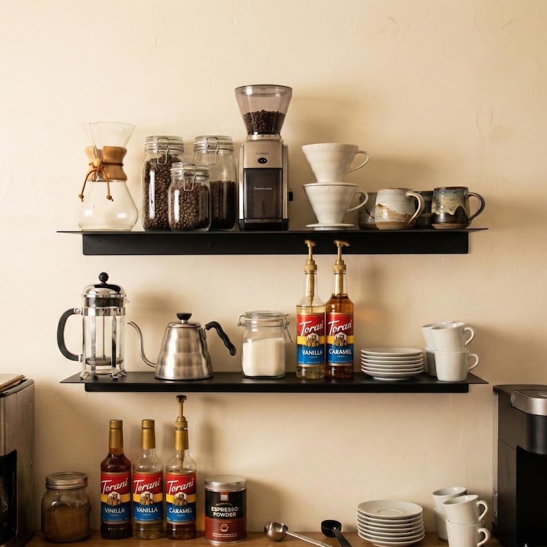 May include: A coffee station with various items on black shelves. Includes a coffee grinder, glass jars with coffee beans, ceramic mugs, a French press, a kettle, Torani vanilla and caramel syrups, and espresso powder.